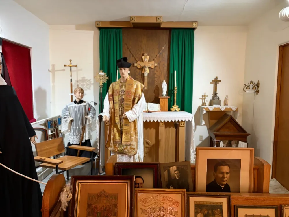 Chapel exhibit with a mannequin priest in gold vestments in front of an altar with a crucifix and green curtains, an altar boy mannequin to the left, and framed portraits of priests in the foreground