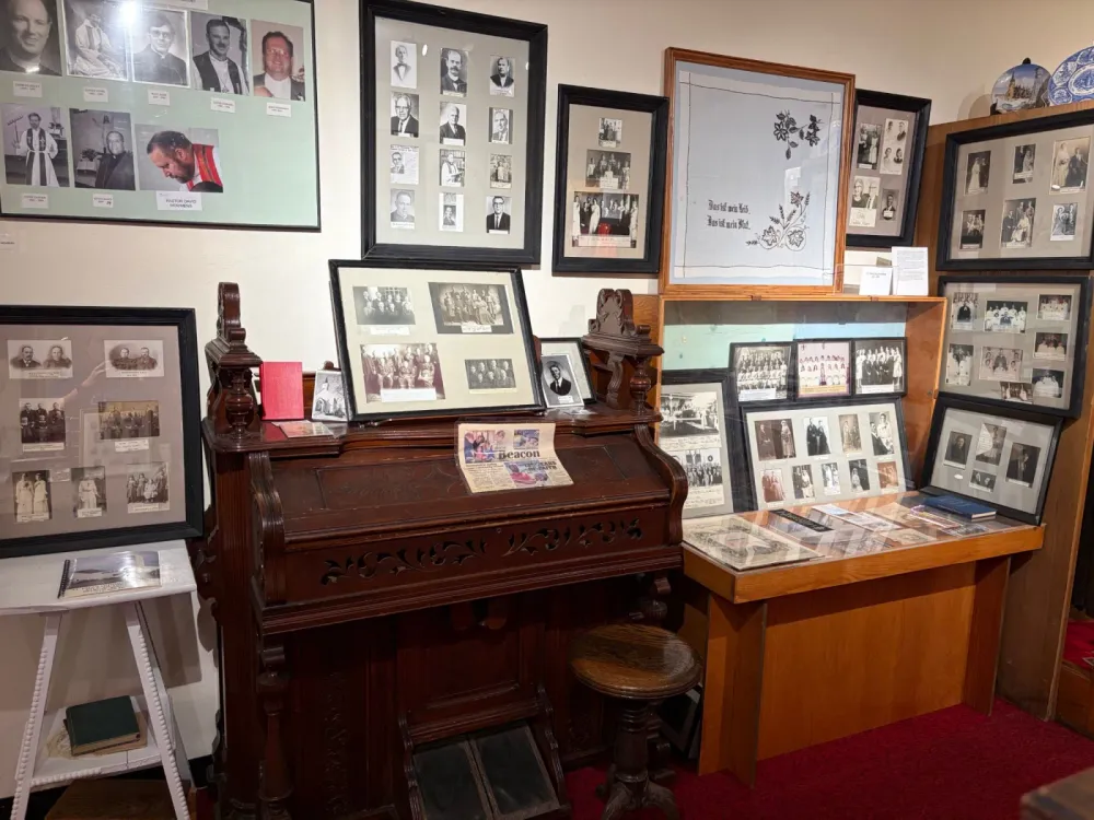 A pump organ surrounded by framed photographs of pastors, church members and congregations, with a glass display case of documents beside it