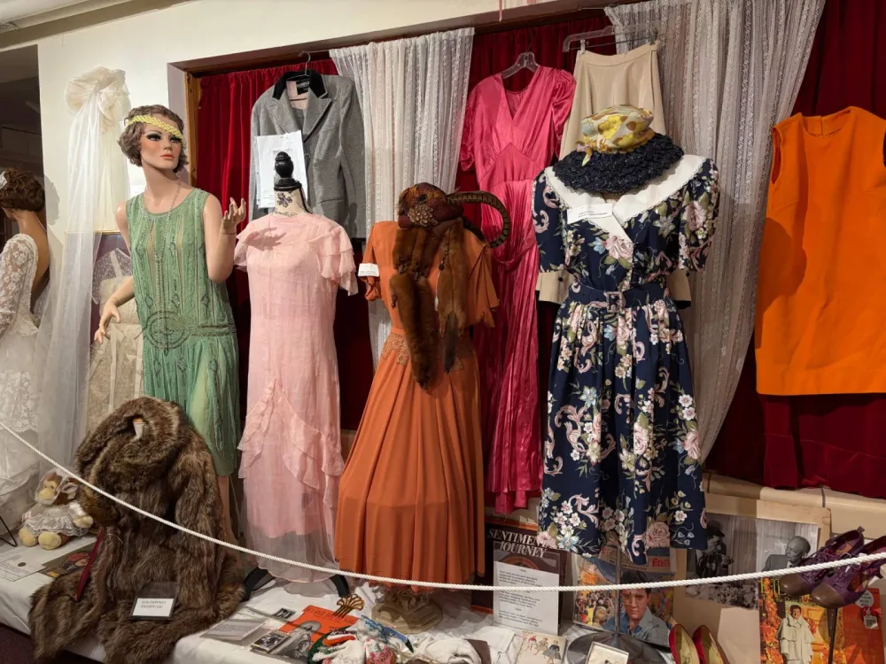 Display of women's dresses from several decades on mannequins and hangers in front of red and lace curtains, including a green beaded dress, a pink chiffon dress, an orange dress with a fur stole and a feathered hat, and a floral dress with a straw hat, with a fur coat on the floor in front
