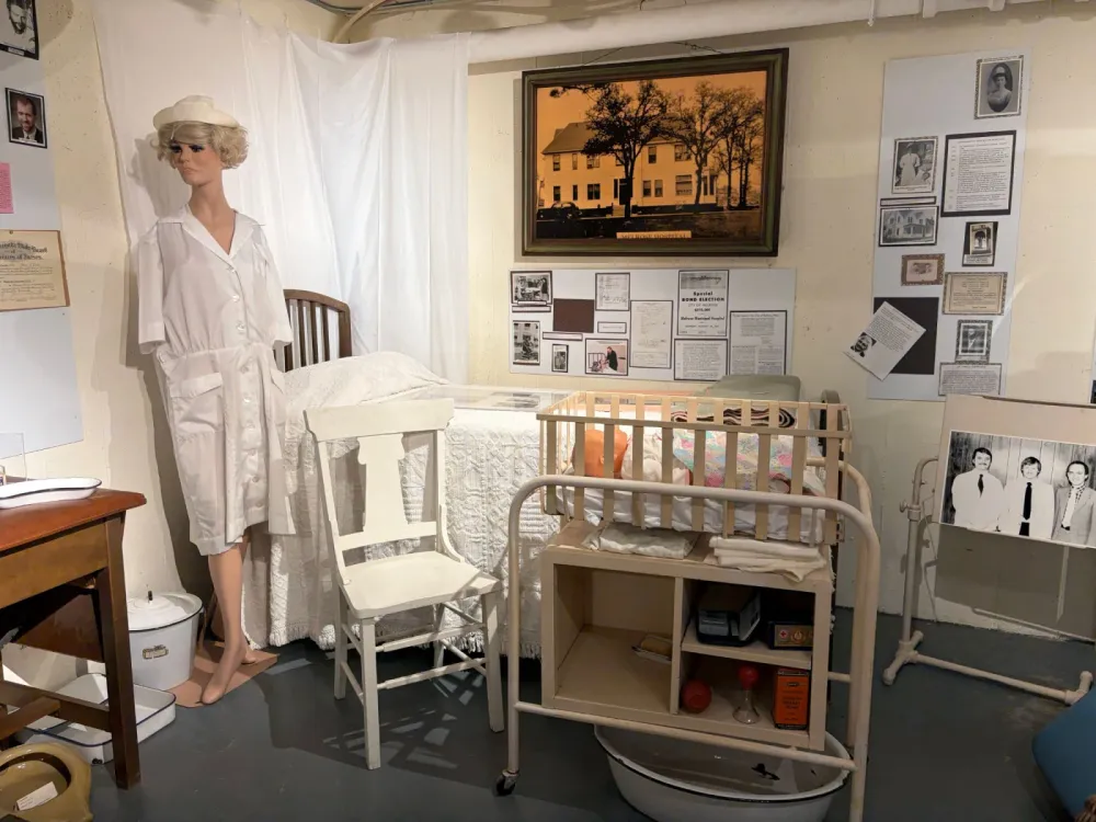 Hospital exhibit in the museum: a mannequin in a white nurse's uniform and cap, a hospital bed, a baby crib on a cart, and wall displays of photographs and documents about the hospital