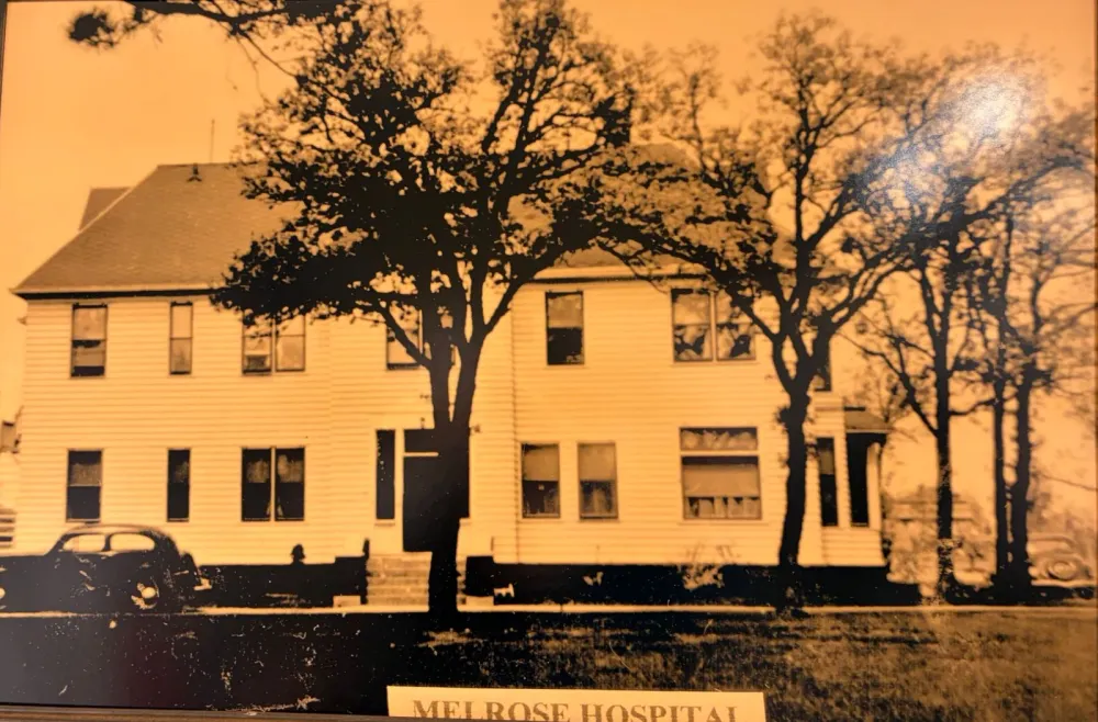 Sepia photograph of the Melrose hospital, a two-story white clapboard building with large trees in front and an old car parked at the left, labeled Melrose Hospital