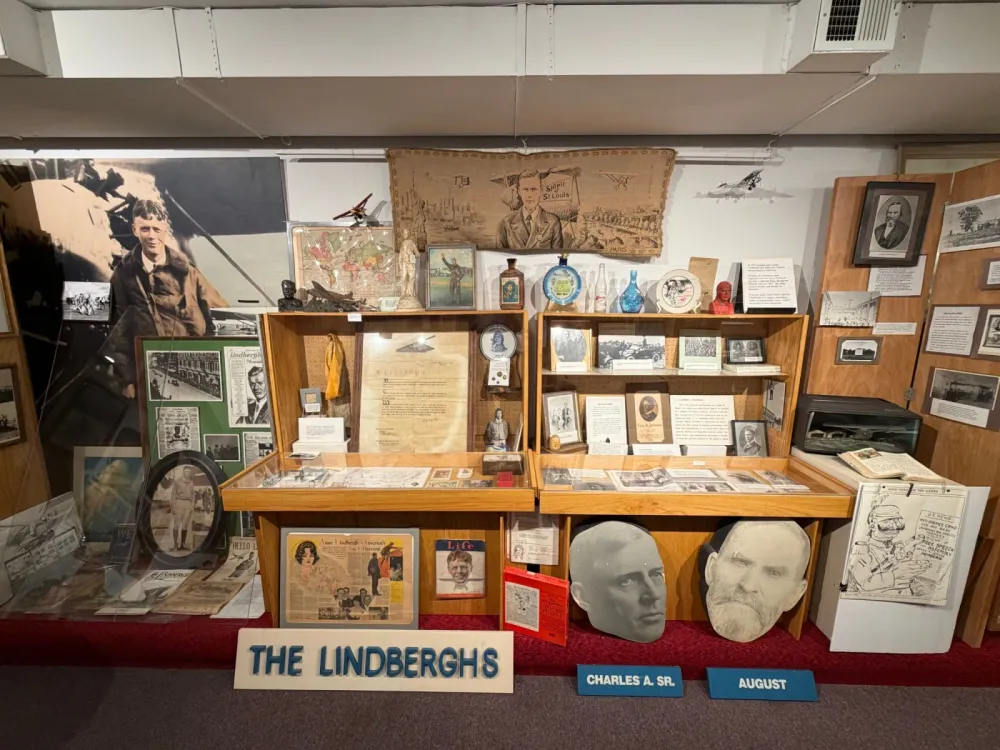The Lindberghs exhibit: display cases and wall displays of photographs, newspapers and memorabilia beneath a Spirit of St. Louis tapestry, with large cutout portraits of Charles A. Sr. and August Lindbergh and a sign reading The Lindberghs