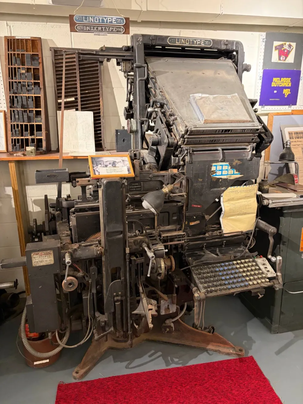 A large black Linotype typesetting machine with a keyboard of brass-colored keys, a slanted magazine of brass molds above it and a hanging Linotype Intertype sign, with a type cabinet and printing supplies behind it