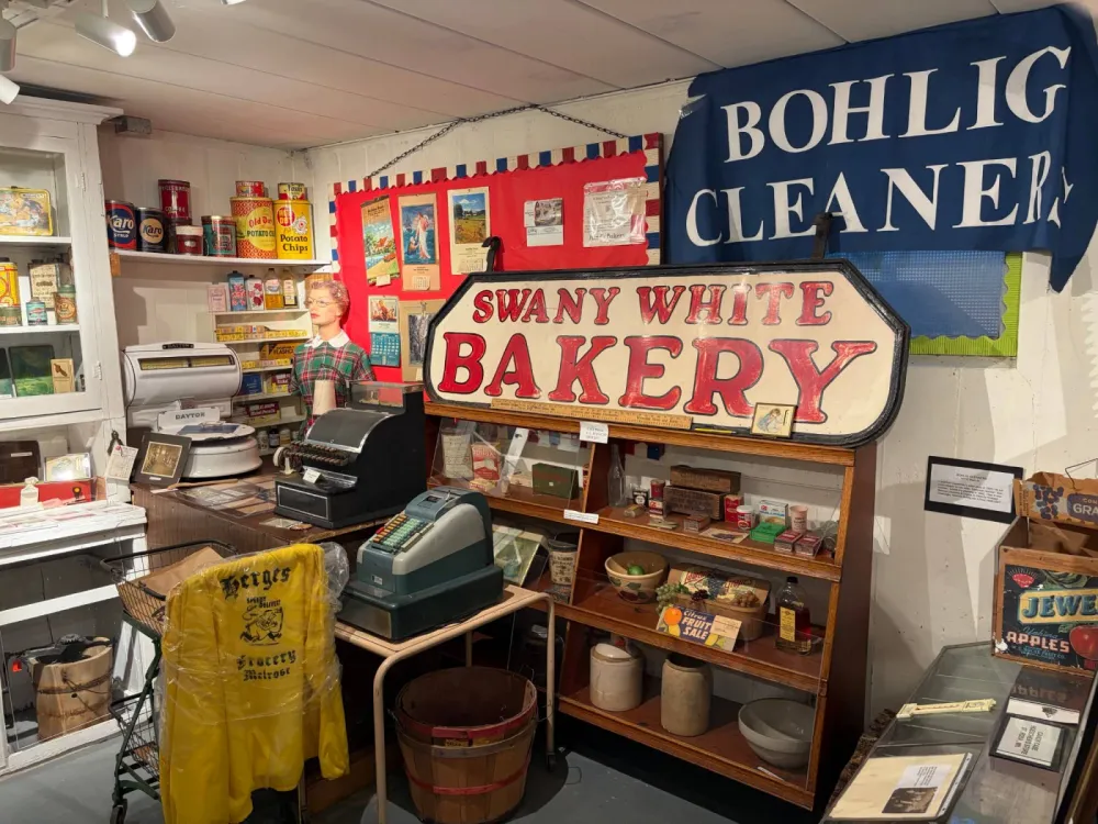 Old-time store display with a hand-painted Swany White Bakery sign, a blue Bohlig Cleaners banner, a mannequin shopkeeper at a Dayton scale and cash registers, shelves of vintage tins and boxes, and a yellow Herges Speedy Delivery Grocery Melrose jacket