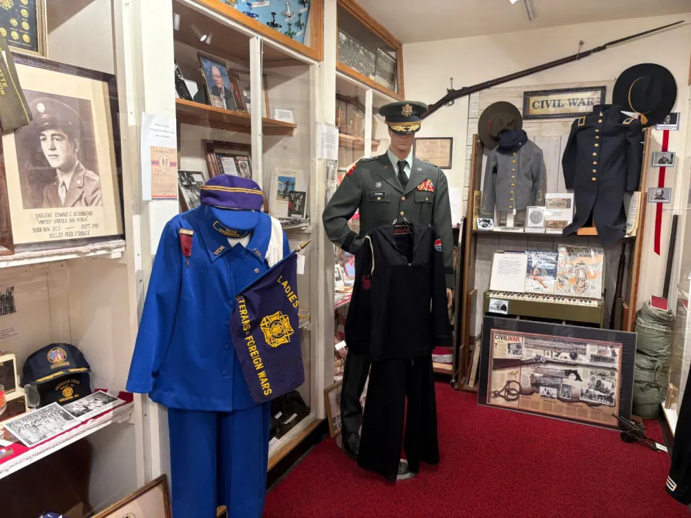 Military exhibit room: a mannequin in a green Army officer's uniform and a blue Veterans of Foreign Wars auxiliary uniform with a flag, a framed portrait of a sergeant killed over France in 1944, and a Civil War corner with Union and Confederate jackets, a rifle and hats