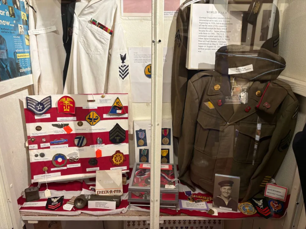 World War II display case with a Navy white uniform, an Army jacket and cap, a board of unit patches and badges, medals and a framed photograph of a sailor