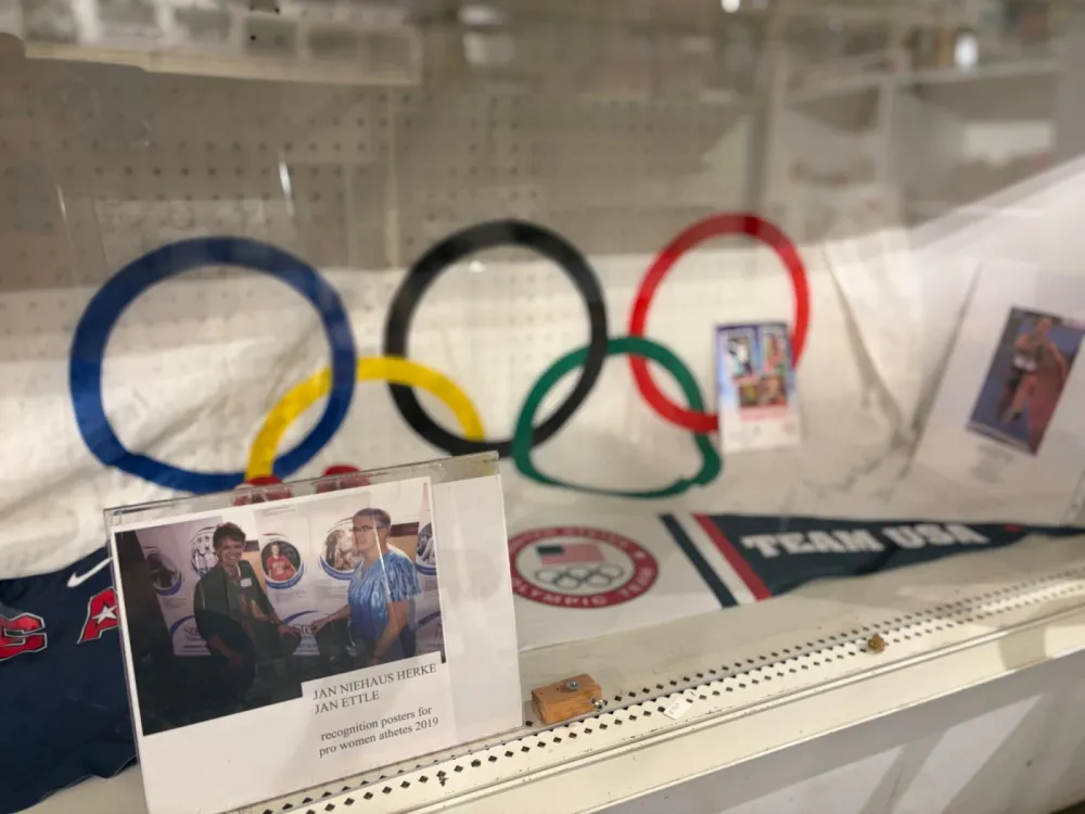 Display case with the five Olympic rings, a Team USA pennant and clothing, an Olympic team emblem, an athlete photo and a framed photo of two women with recognition posters