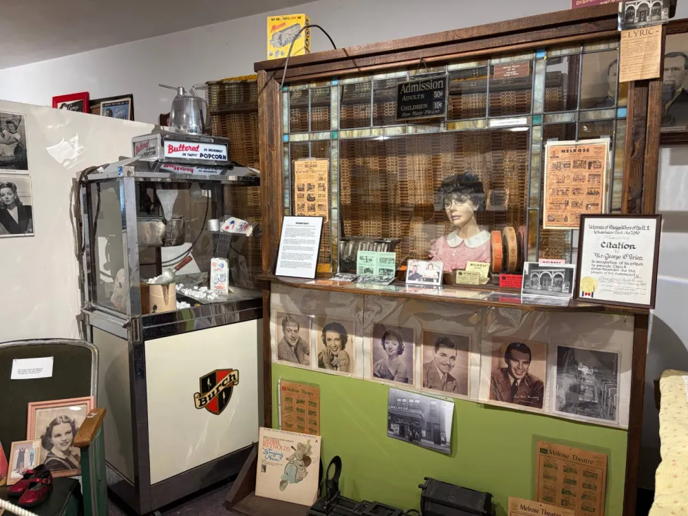 Melrose Theater exhibit: a wooden ticket booth with a mannequin ticket seller and an Admission sign reading Adults 50 cents, Children 10 cents, beside a chrome popcorn machine, with movie posters and portraits of film stars on the wall