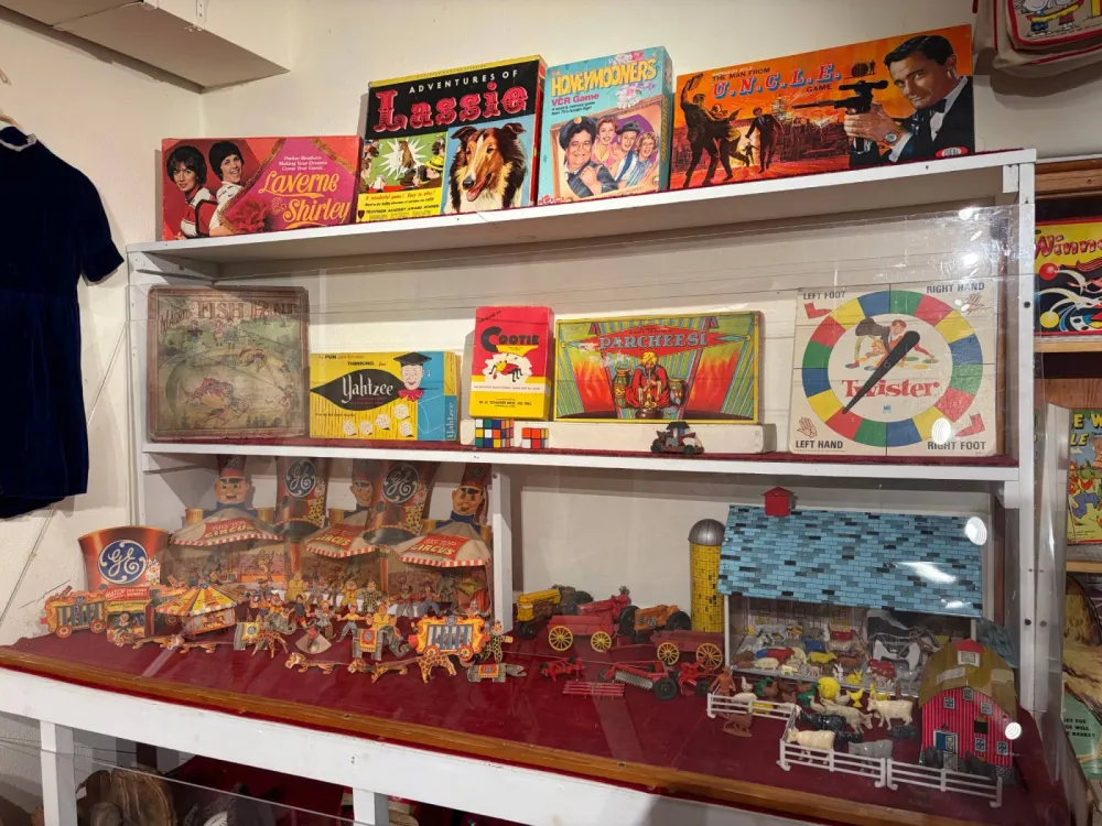 Shelves of vintage board games and toys: Laverne & Shirley, Lassie, The Honeymooners and Man from U.N.C.L.E. games, Magnetic Fish Pond, Yahtzee, Cootie, Parcheesi and Twister, with paper circus cutouts, toy tractors and a tin barn set below