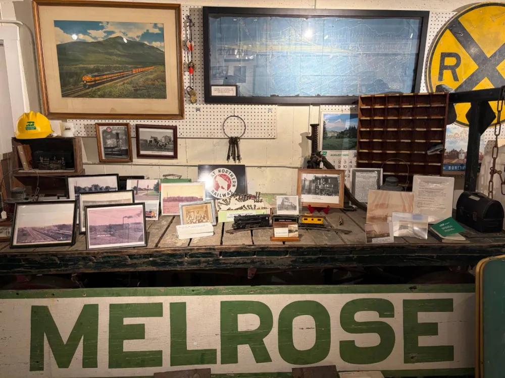 Railroad display on a worn wooden freight cart: a painted green and white MELROSE station sign, framed photographs of the Melrose rail yards and roundhouse, a Great Northern Railway emblem, a Burlington Northern hard hat, a toy locomotive, lanterns, a telegraph key, a railroad crossing sign and a framed map